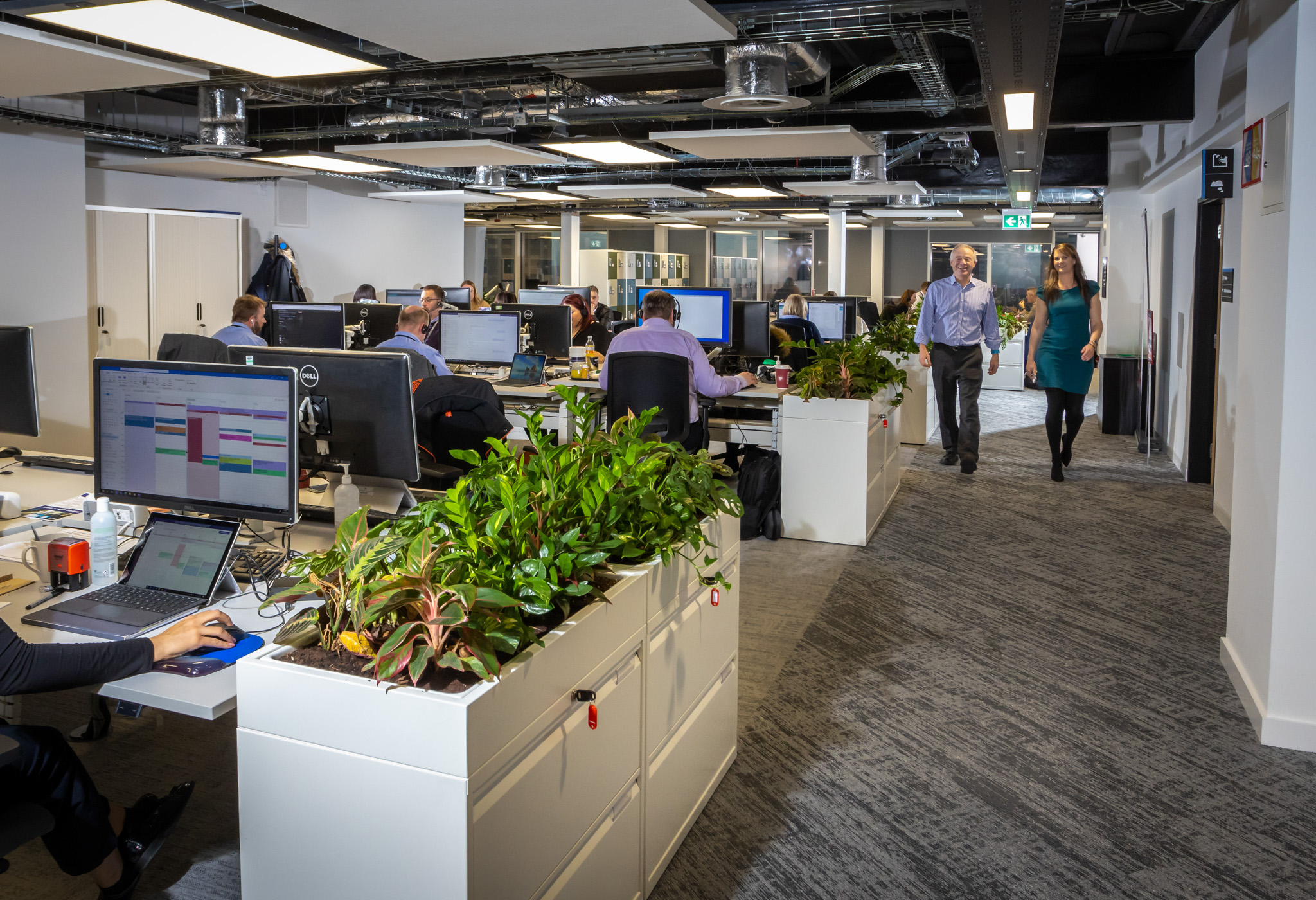 internal office, rows of desk with people working on computers. two people walking down gangway.